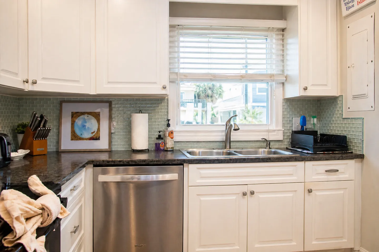 Kitchen counter with stainless steel double sink, dishwasher, dark granite countertop, and white shaker cabinets under a window with blinds.