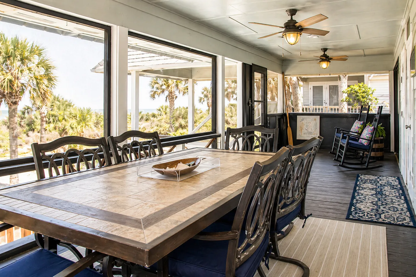 Screened porch with a large dining table and chairs, ceiling fans, and rocking chairs overlooking coastal palm trees.