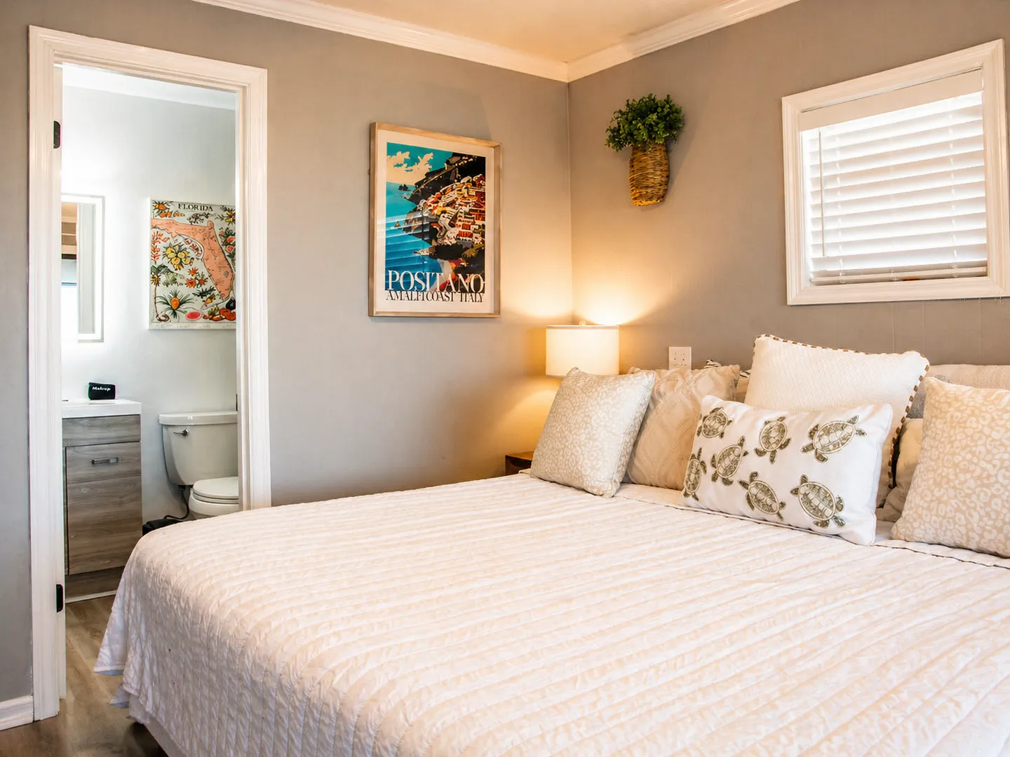 Bedroom with a made bed, accent pillows, framed art, and an open doorway leading to an en-suite bathroom.