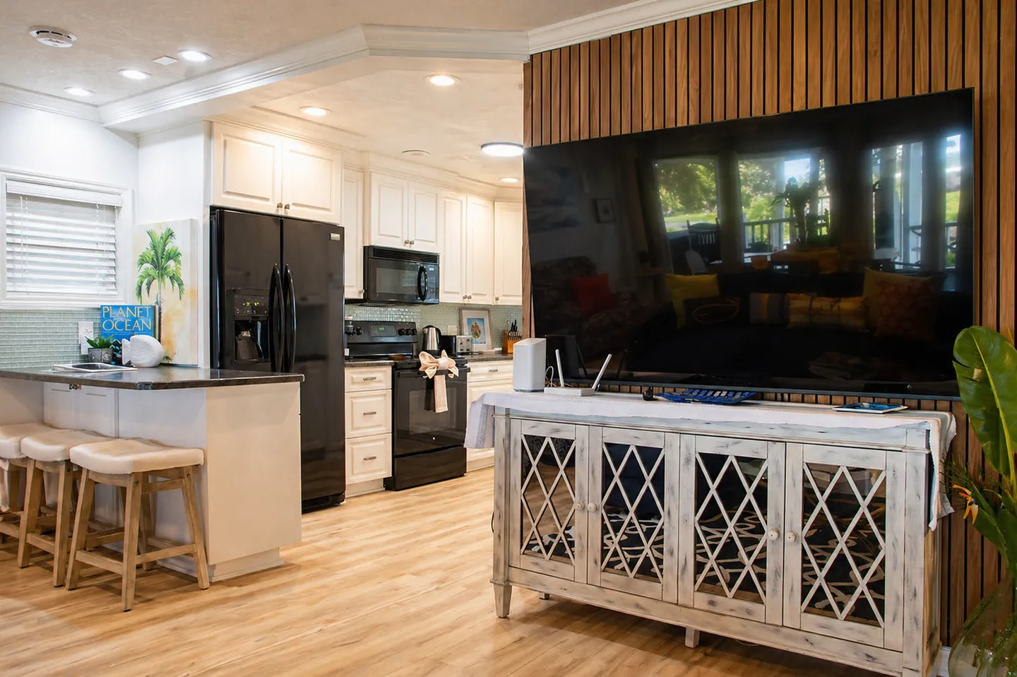 Open-concept living and kitchen area featuring a large wall-mounted television over a white credenza next to a kitchen island with barstools.