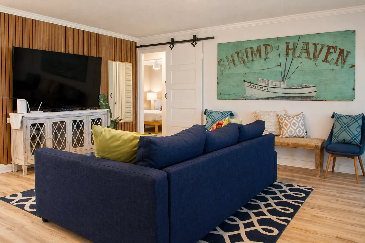 Living room featuring a deep blue sectional sofa, a wall-mounted TV over a white credenza with a wood slat accent wall, and coastal wall art.