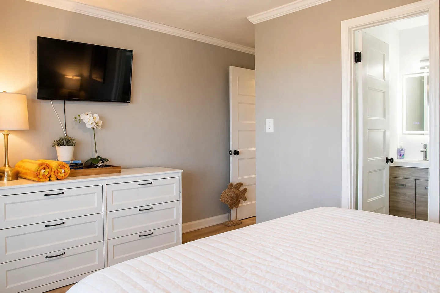 Bedroom with a white dresser, a wall-mounted flat-screen TV, a corner of a bed, and an open doorway leading to a bathroom.