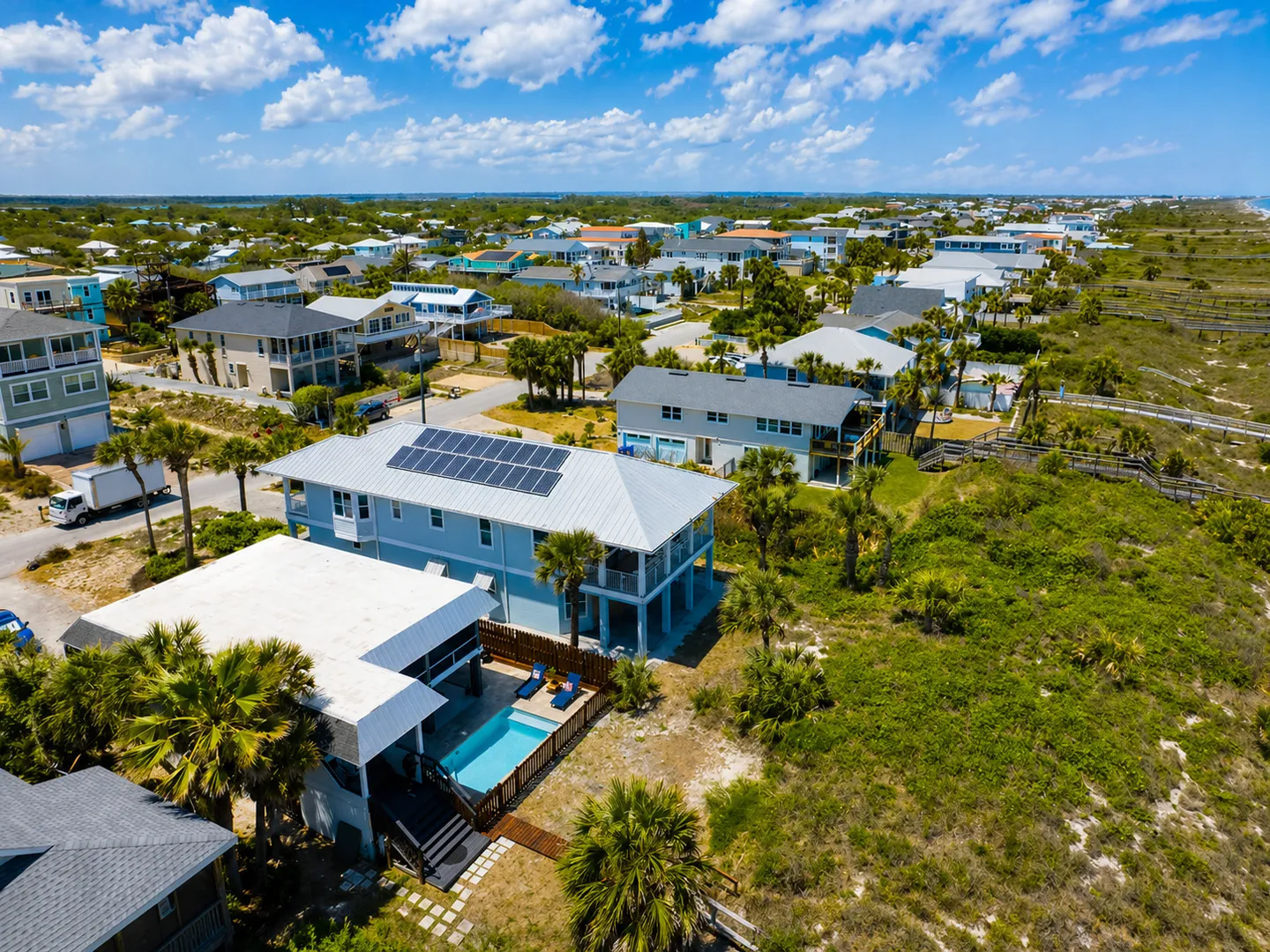 Aerial view of a coastal neighborhood showing a two-story blue beach house with solar panels and a private backyard pool next to vegetated dunes.