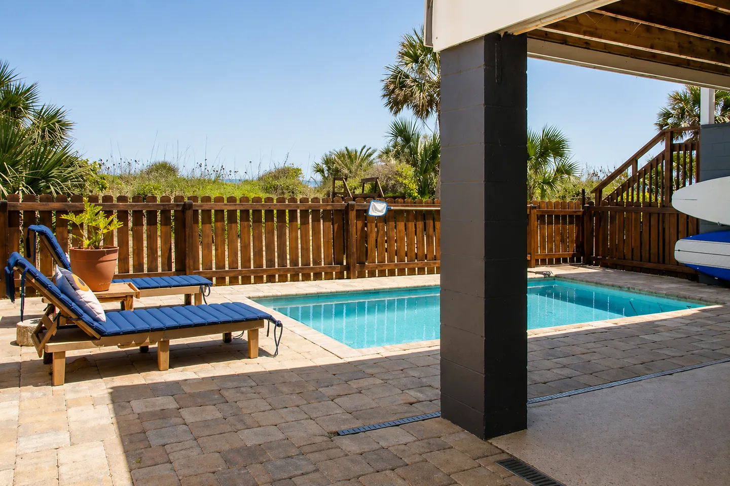 Outdoor paved patio area featuring a private swimming pool, cushioned lounge chairs, and a wooden fence overlooking coastal vegetation.