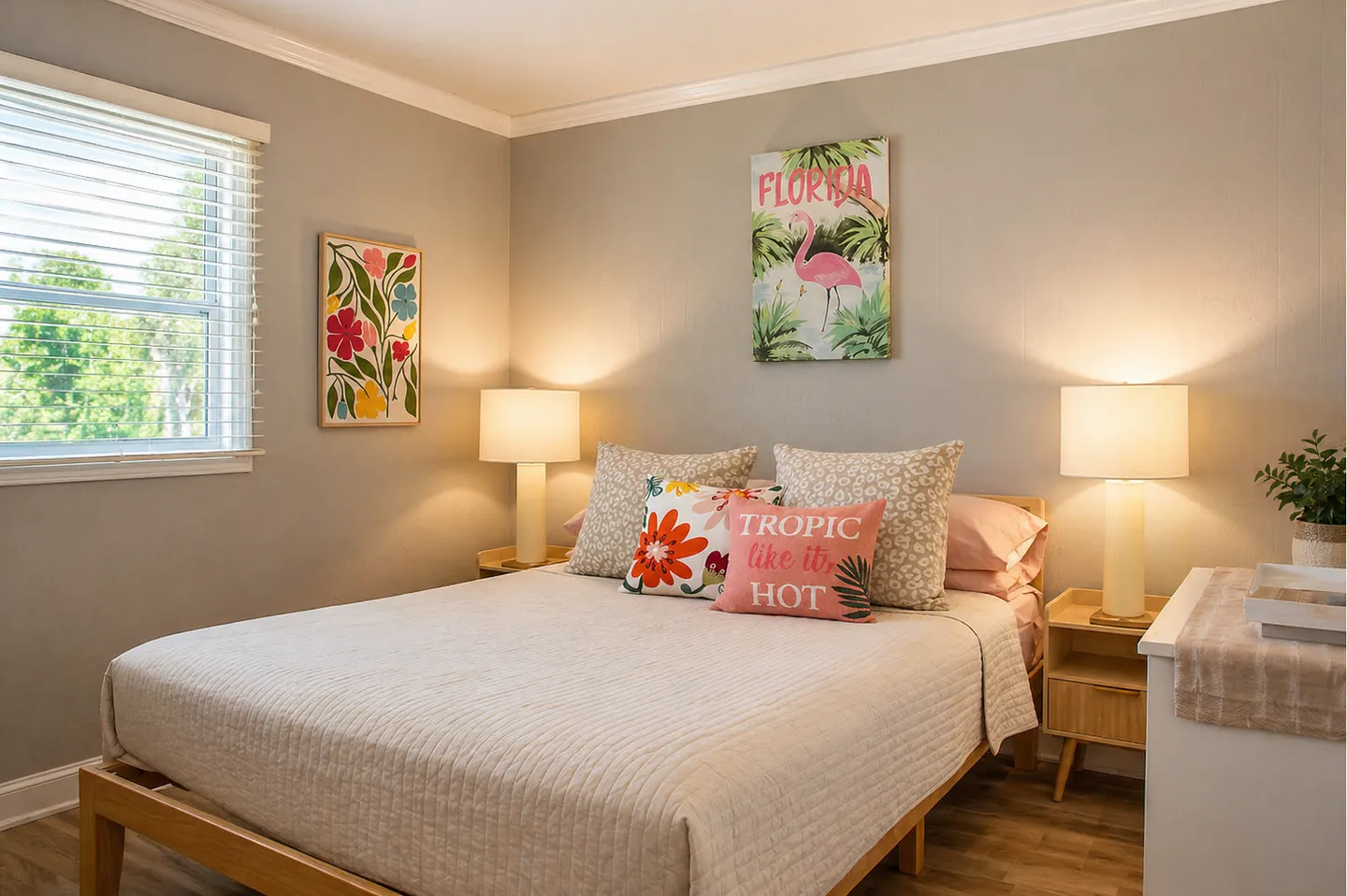 Bedroom with a wooden platform bed flanked by two nightstands with glowing table lamps, tropical wall art, and a window with blinds.