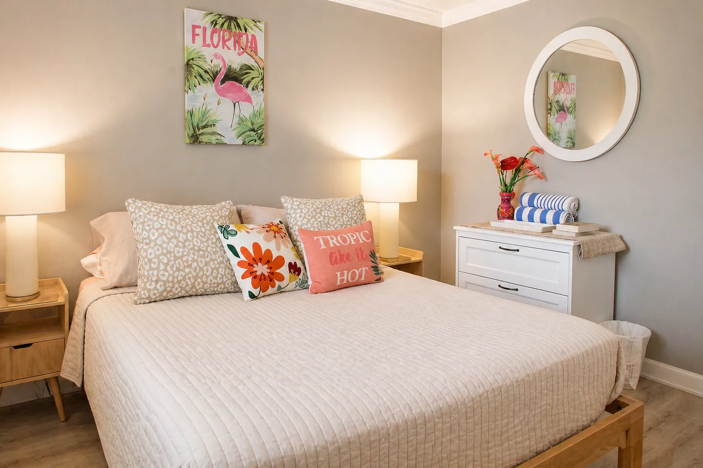 Bedroom with a neatly made bed flanked by nightstands with glowing table lamps, a flamingo wall art print, and a white dresser beneath a round mirror.