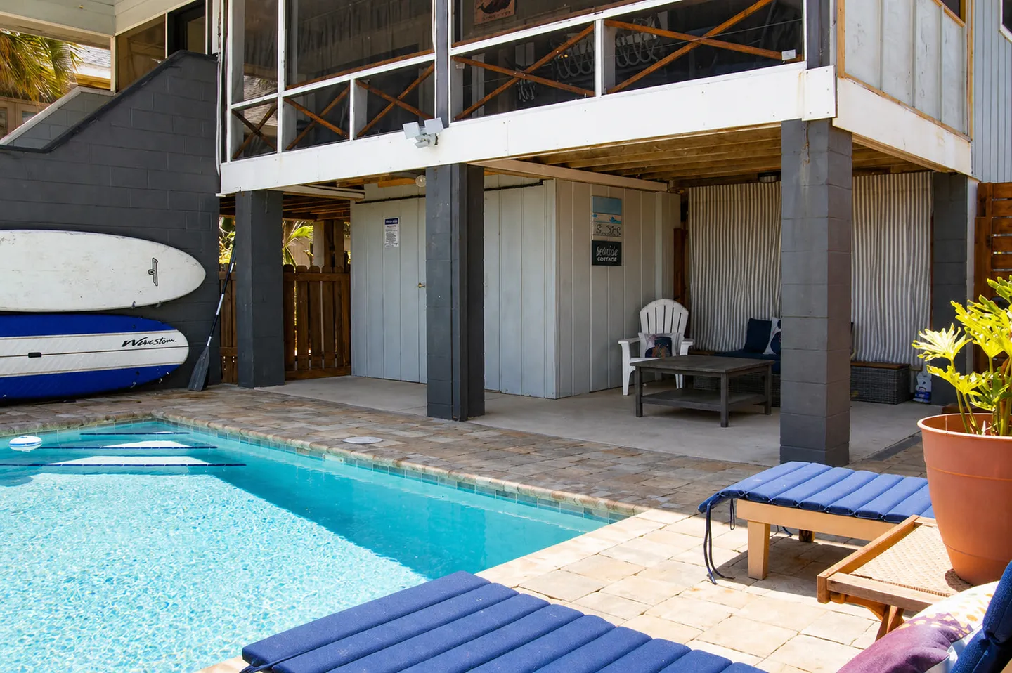 Swimming pool patio with cushioned lounge chairs next to a covered lower level with outdoor seating and stored surfboards.