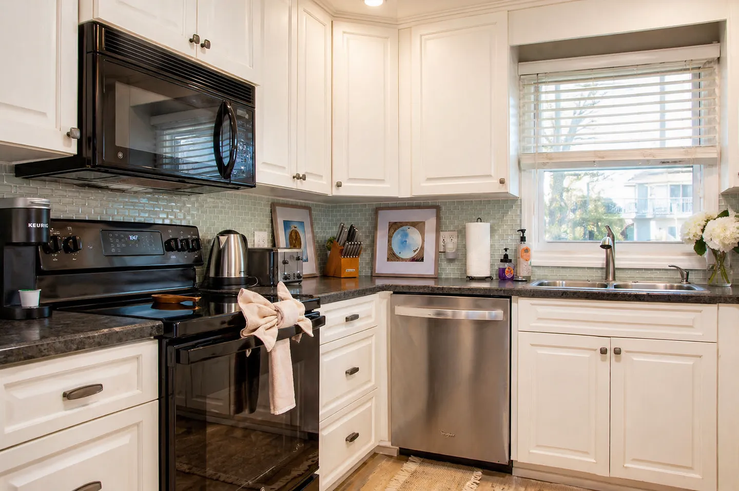 Kitchen corner showing white cabinets, dark countertops, a black range with over-the-range microwave, stainless steel dishwasher, and a window above the sink.