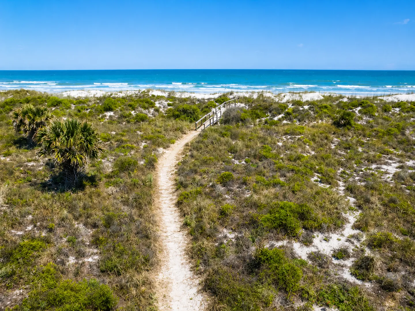 Sandy path leading through coastal dunes and vegetation toward a wooden boardwalk and the ocean.