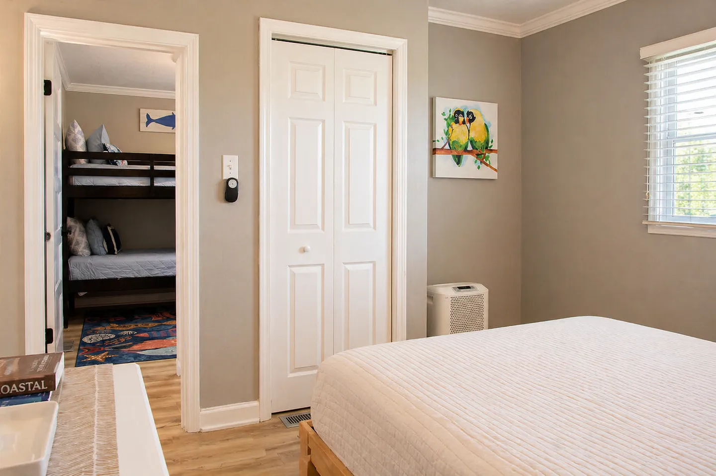 Bedroom with a white-dressed bed, closet door, parrot art, and a view into an adjacent bunk bed room.