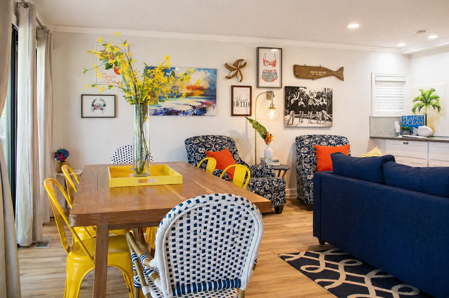 Dining and living space with a wooden dining table, colorful chairs, floral armchairs, blue sofa, and beach-themed wall art.