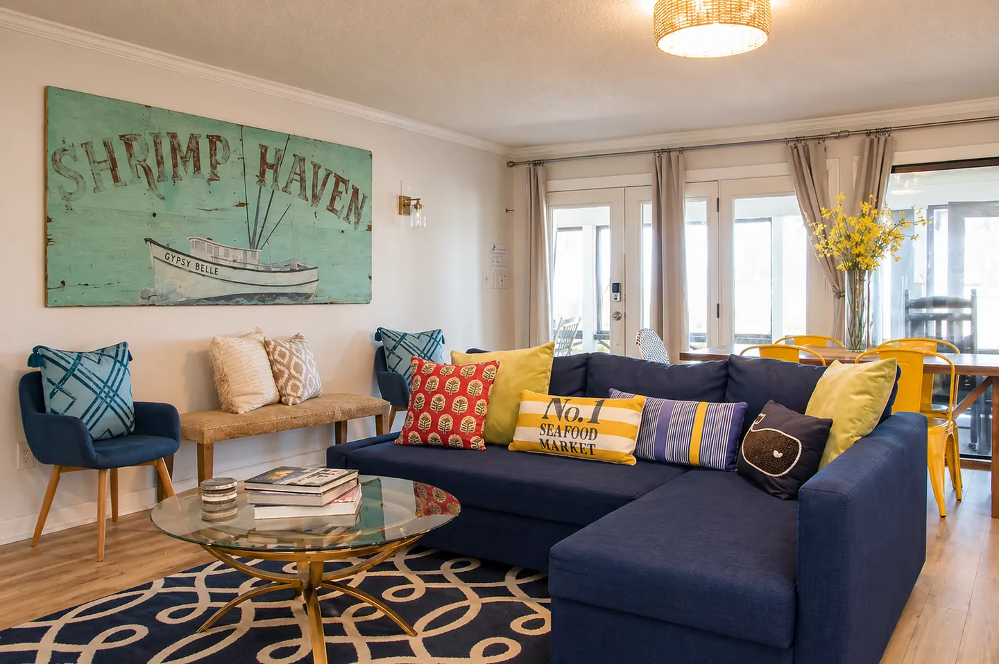 Living room with a large blue sectional sofa, round glass coffee table, patterned rug, and nautical-themed wall art.