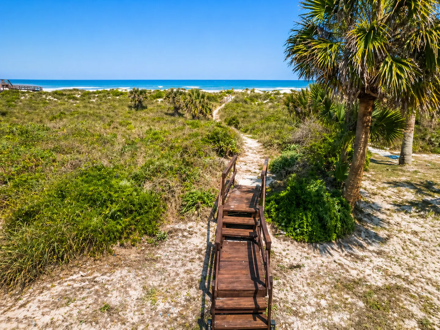 Elevated view of wooden stairs leading to a sandy footpath through coastal dunes and vegetation toward the ocean under a clear blue sky.