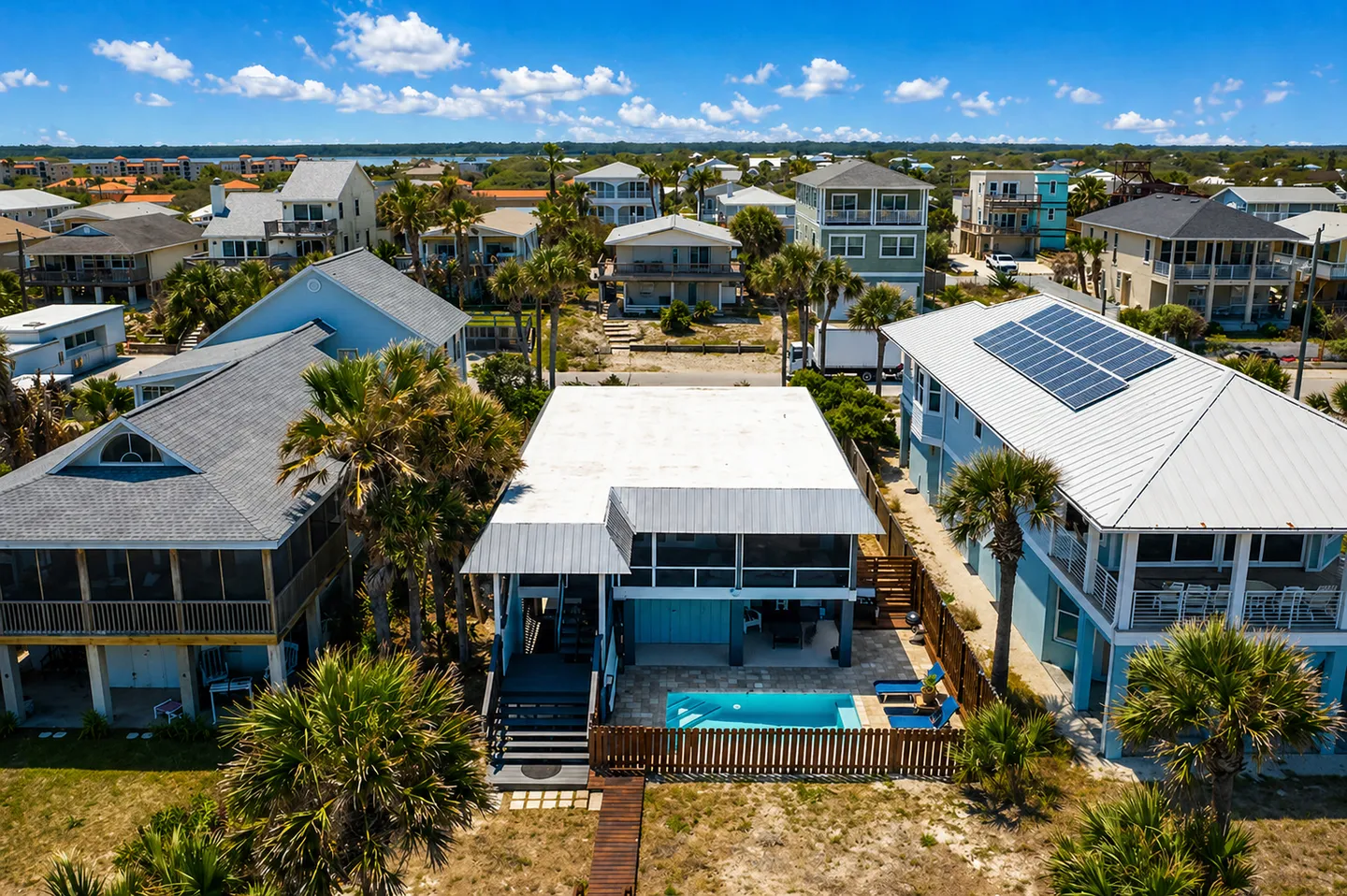 Aerial view of a coastal beach house featuring a private swimming pool, fenced patio, and surrounding neighborhood under a blue sky.