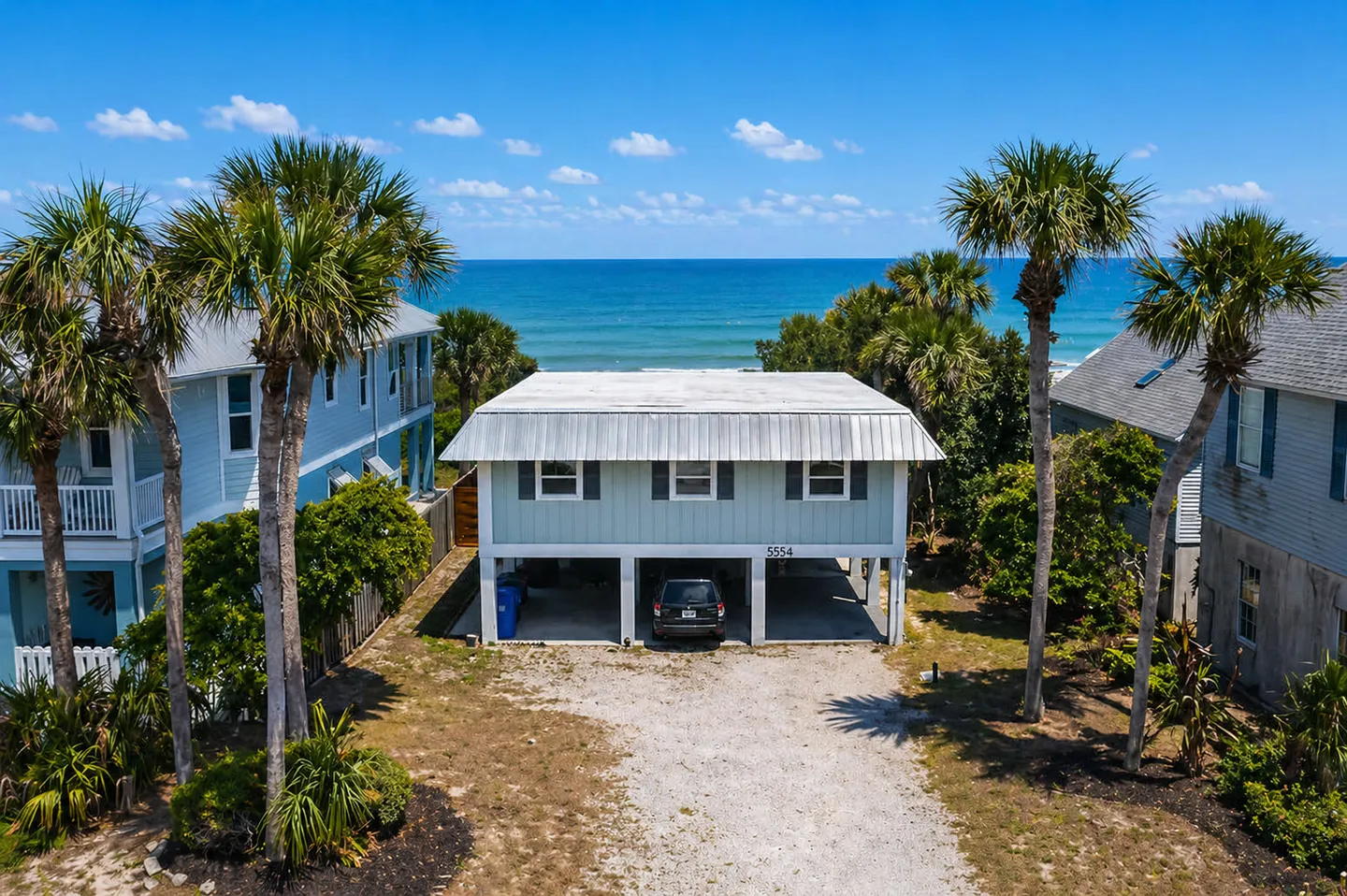 Elevated view of a stilt beach house with car parked beneath, palm trees, and ocean in the background