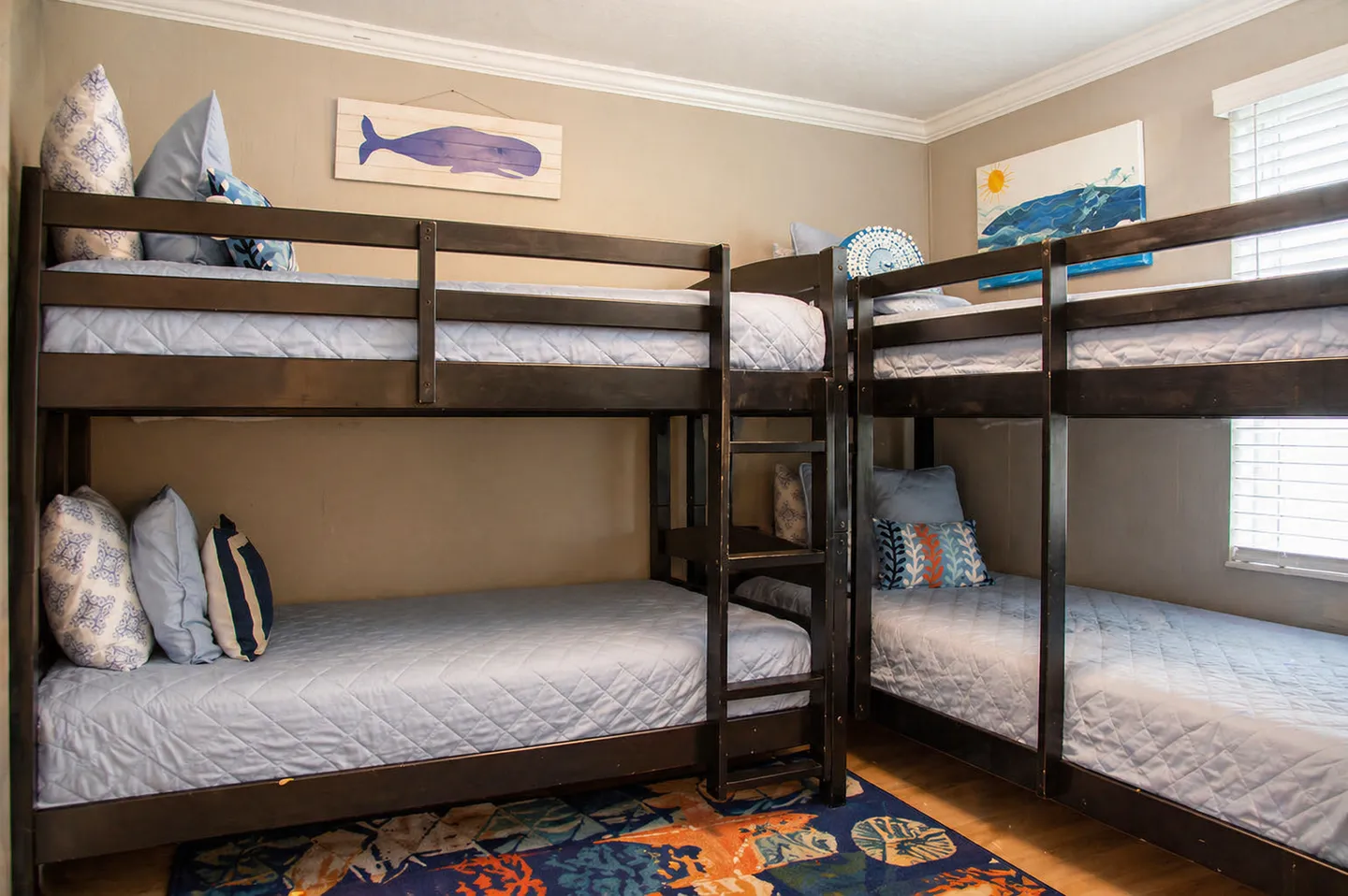 A bedroom corner featuring two dark wood bunk bed sets arranged in an L-shape with light blue quilted bedding and ocean-themed wall art.
