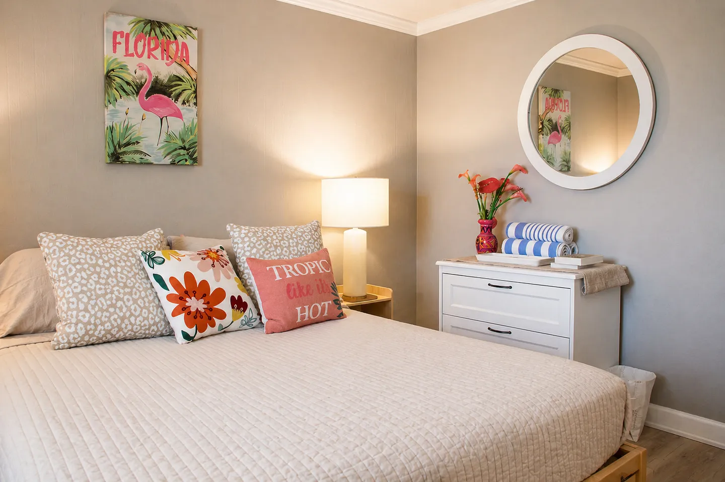 Bedroom with a made bed, Florida flamingo artwork, a nightstand with a lamp, and a white dresser beneath a round mirror.