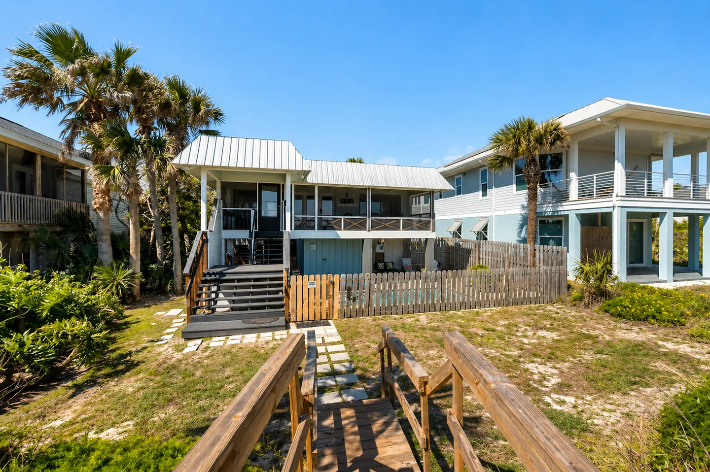 Exterior view of a coastal home with a metal roof, fenced yard, swimming pool, and wooden walkway stairs under a clear blue sky.