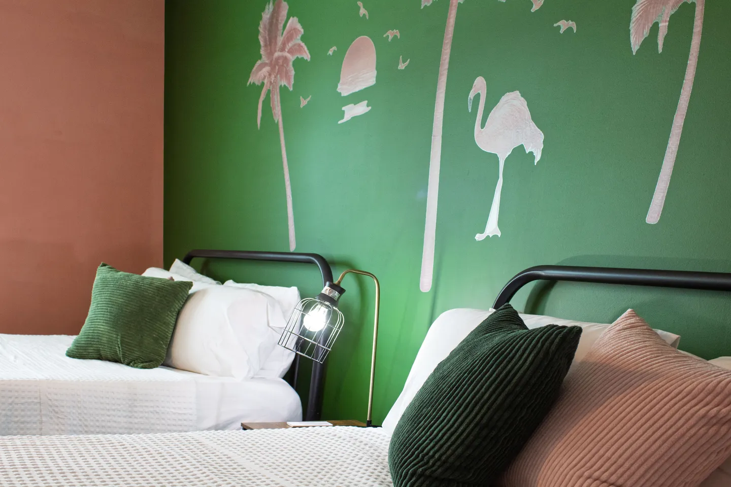 Close-up of two beds with dark metal headboards, green and pink accent pillows, and a green accent wall with painted tropical flamingo and palm tree silhouettes.