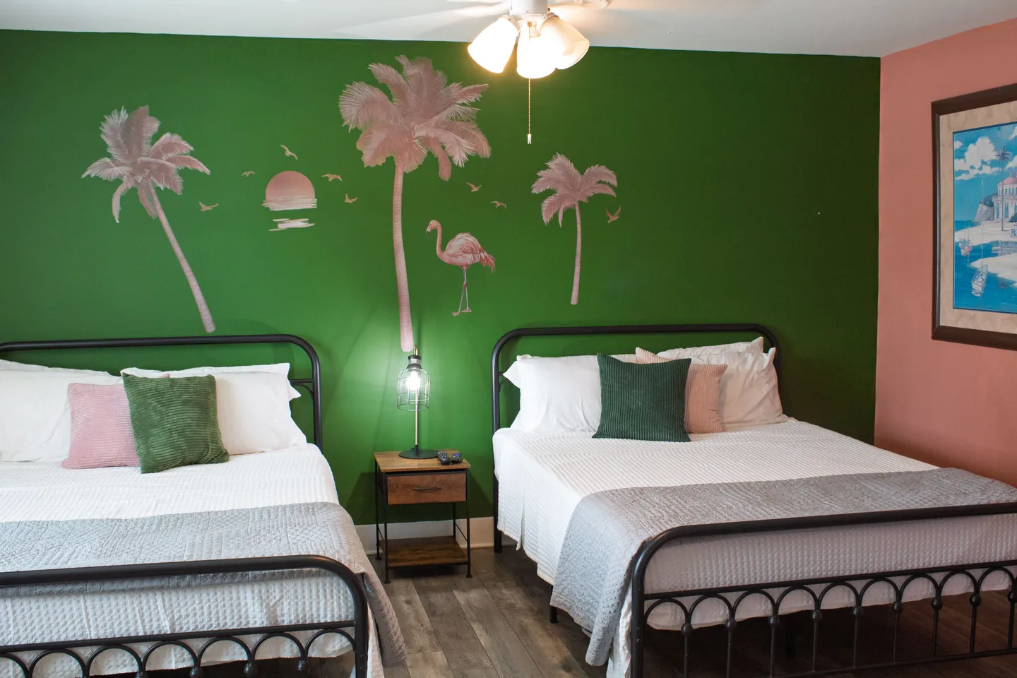 A bedroom with two beds featuring black metal frames against a green accent wall with painted palm trees and a flamingo.