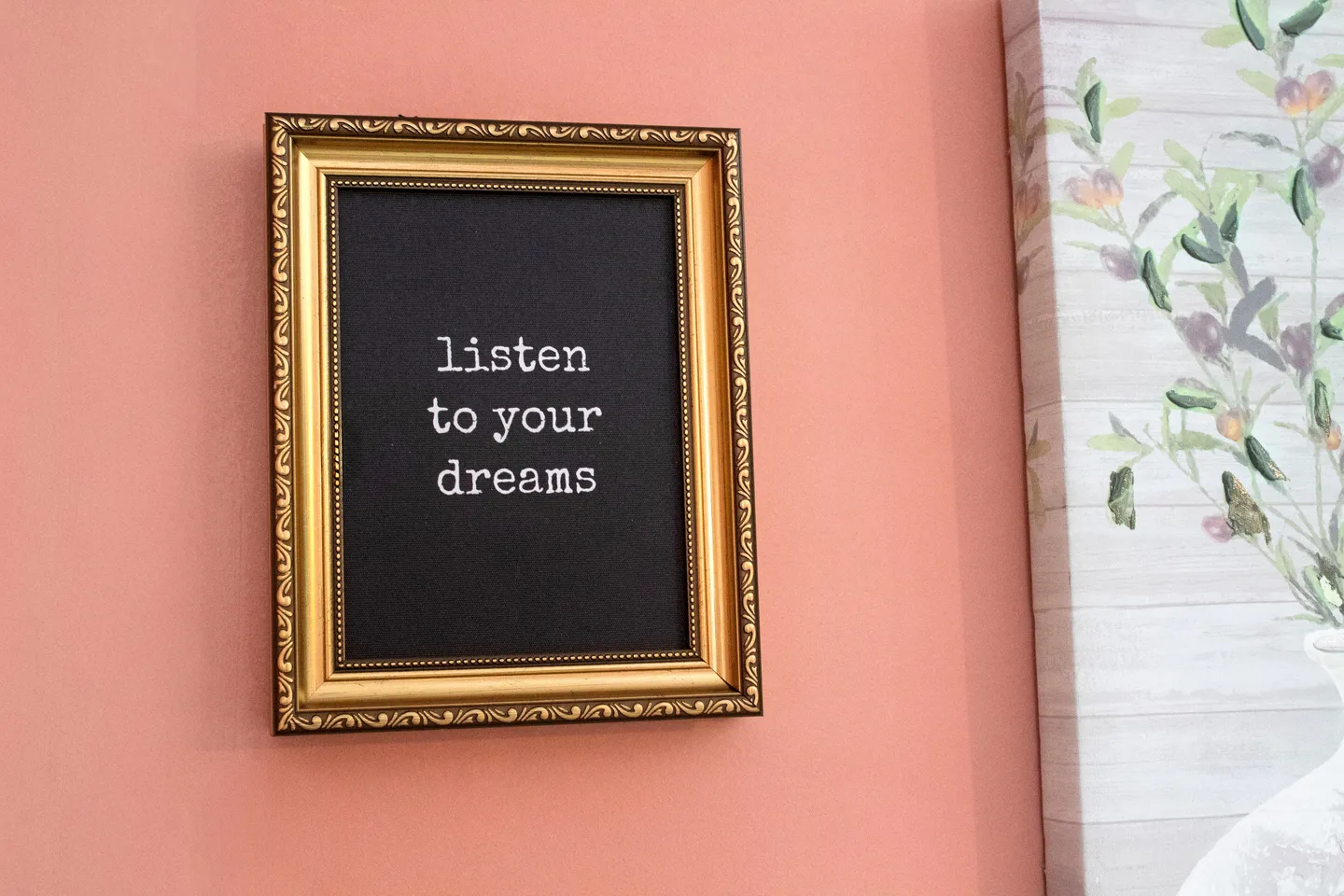 A framed quote reading 'listen to your dreams' hangs against a coral-pink wall next to canvas artwork.