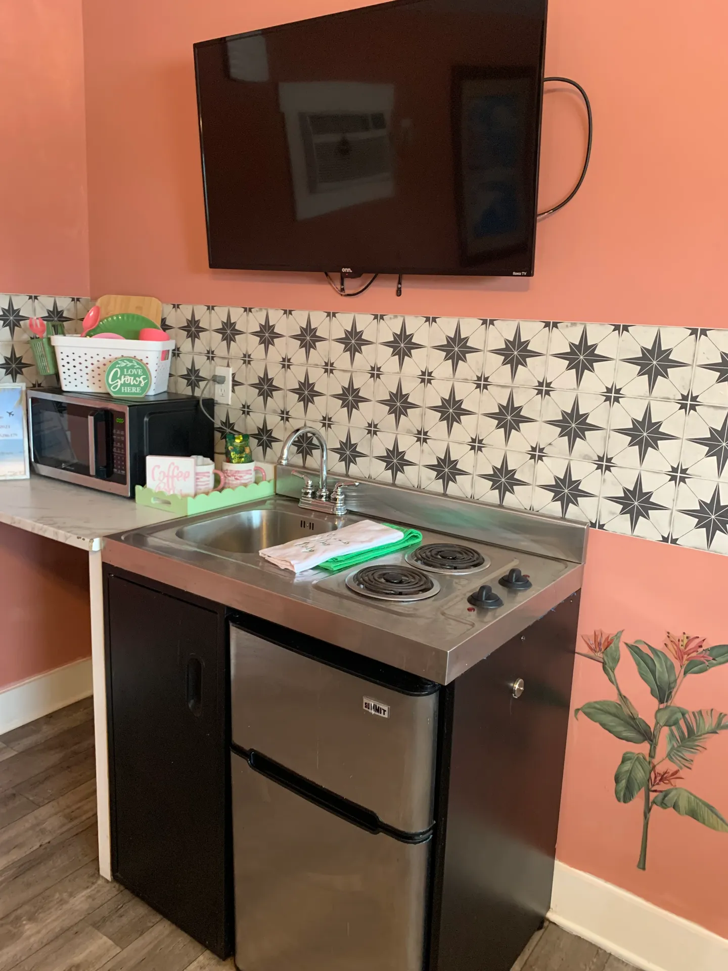 A compact kitchenette setup featuring a stainless steel combo sink and two-burner stovetop over a mini fridge, alongside a microwave and wall-mounted television against pink walls.