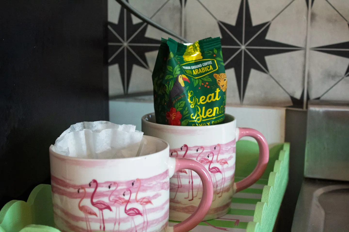 Close-up of flamingo-themed coffee mugs with coffee filters and a bag of coffee on a green tray.