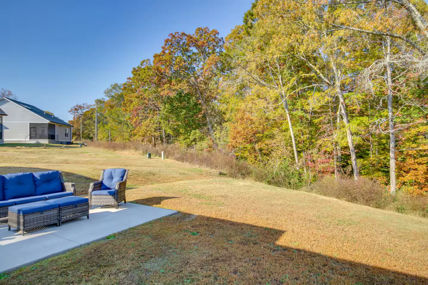A concrete patio with blue outdoor seating looks out over a grassy yard bordered by autumn trees under a clear blue sky.