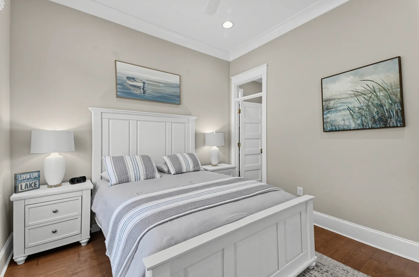 Bedroom featuring a white wooden panel bed, matching nightstands with lamps, coastal artwork, and neutral walls.
