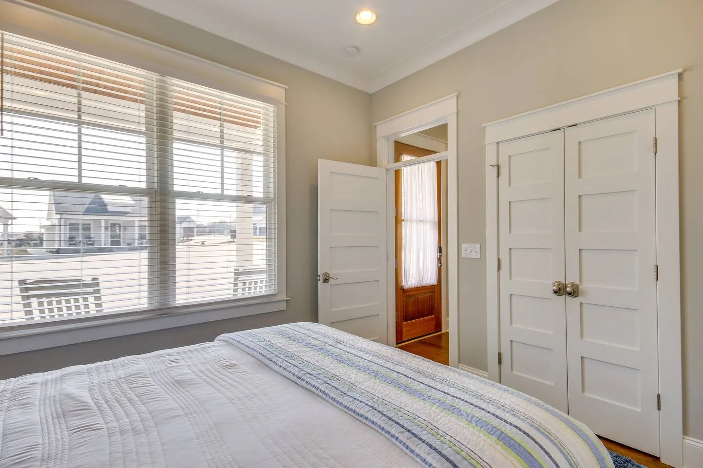 Bedroom corner showing a bed with white bedding, a large window with blinds, and white interior doors.