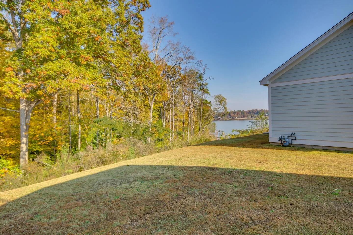 A sloping grassy backyard next to the side of a house, overlooking autumn trees and a lake.