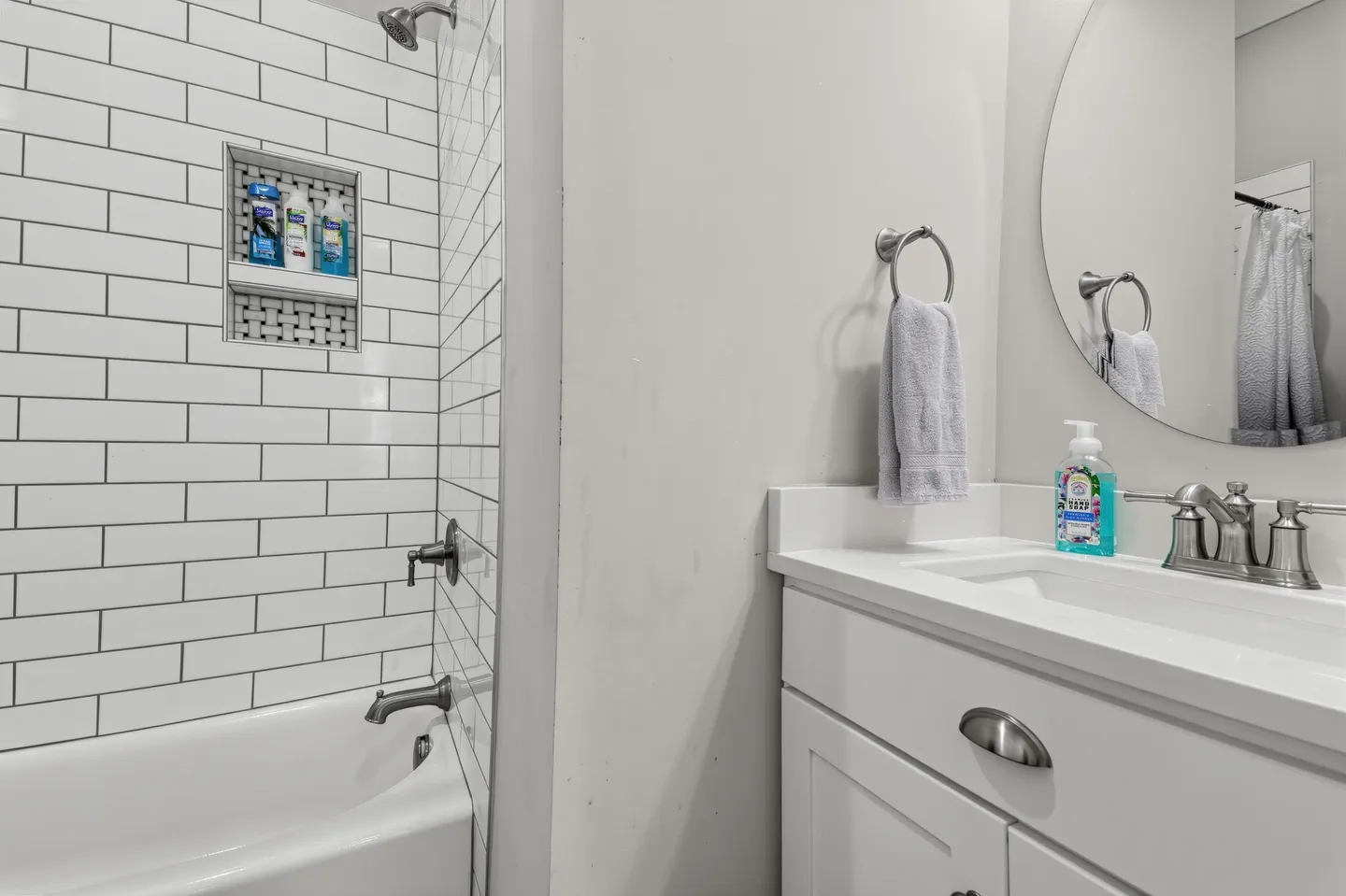 Bathroom showing white subway tile bathtub shower combination next to a white single vanity with a round mirror.