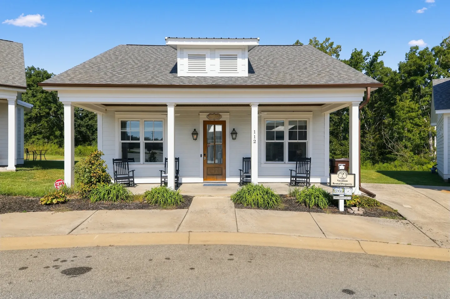 Front exterior of a white single-story cottage with a covered front porch featuring rocking chairs.