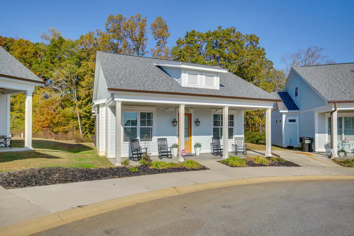 Exterior front view of a small, white single-story cottage with a covered front porch featuring rocking chairs under a blue sky.