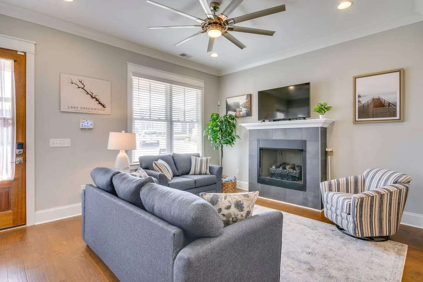 Living room with gray upholstered sofas, a striped armchair, a tiled fireplace with a mounted TV, and a large ceiling fan.