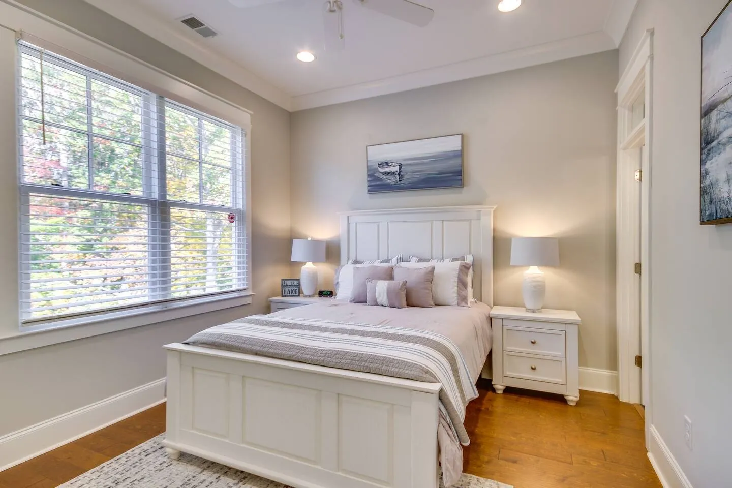 Bedroom featuring a white wooden bed flanked by matching nightstands with lamps and a large window with horizontal blinds.