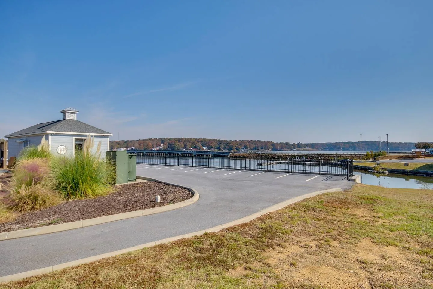 A paved driveway and parking area bordered by a black metal fence, overlooking a body of water with distant docks under a clear blue sky.