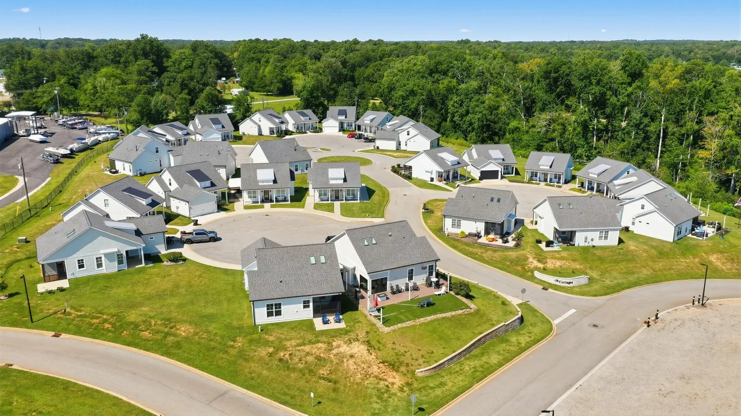 Aerial view of a suburban neighborhood cul-de-sac with white, gray-roofed single-story cottages surrounded by green lawns and trees.