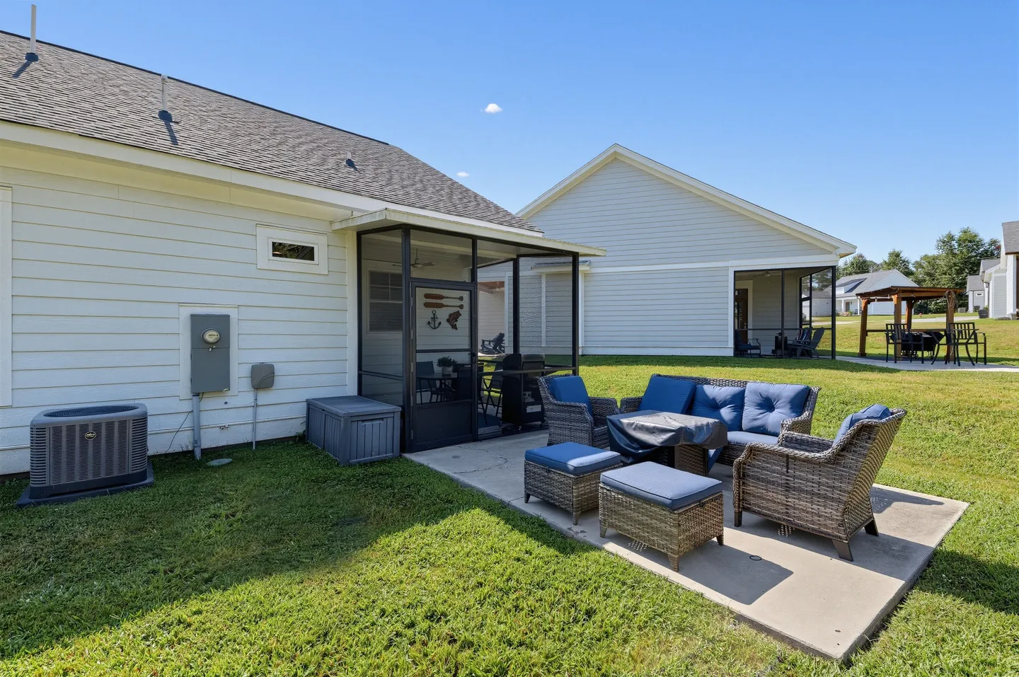 Backyard patio with wicker seating and cushions outside a screened porch, next to a grassy lawn under a clear sky.