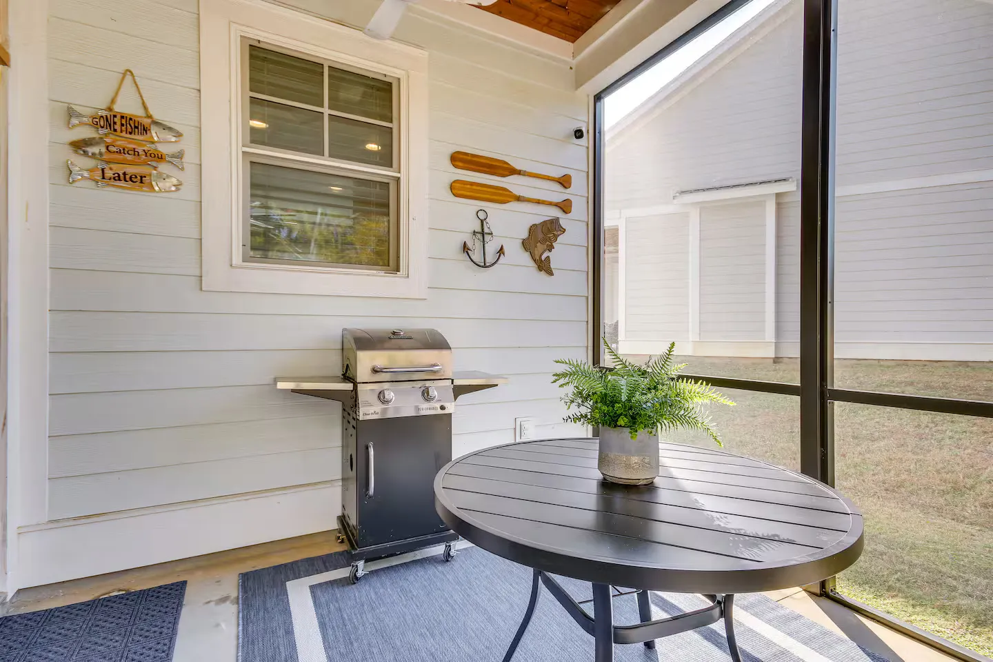Screened porch with a freestanding barbecue grill, round outdoor table with a potted plant, and nautical wall decor.