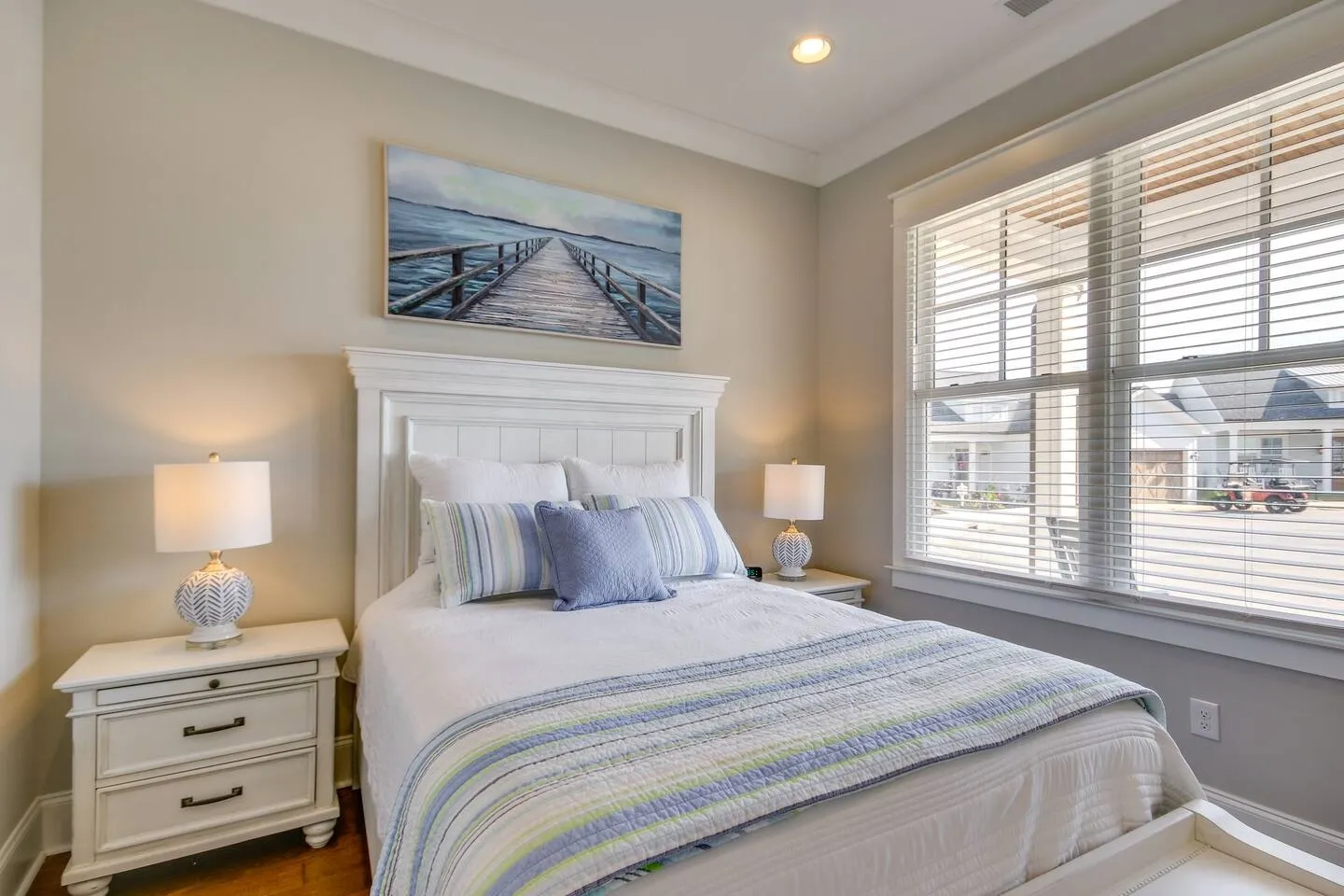 Bedroom with a white headboard, blue and white bedding, two matching white nightstands with lamps, and a window with horizontal blinds.
