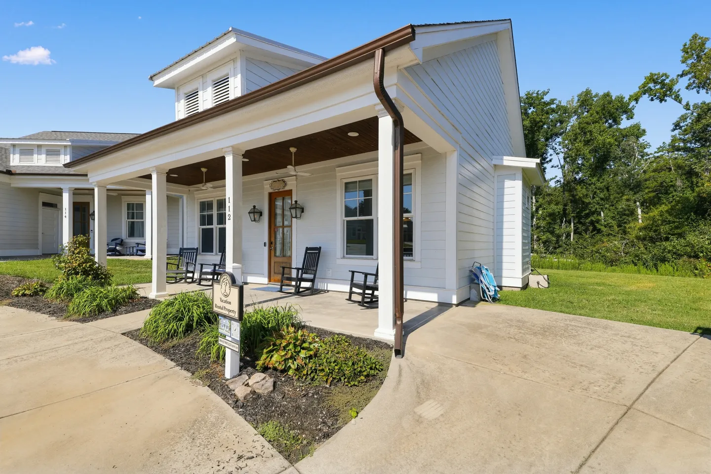 Front porch of a white cottage with rocking chairs, columns, and a concrete driveway on a sunny day.