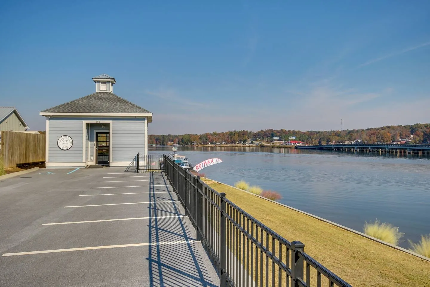 A waterfront parking area and small blue building with a cupola beside a river, bordered by a black metal fence.