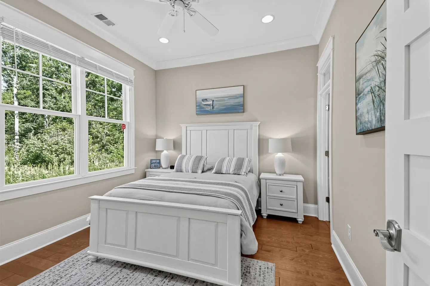 Bedroom with a white panel bed flanked by matching nightstands, hardwood flooring, and a large window showing green foliage outside.