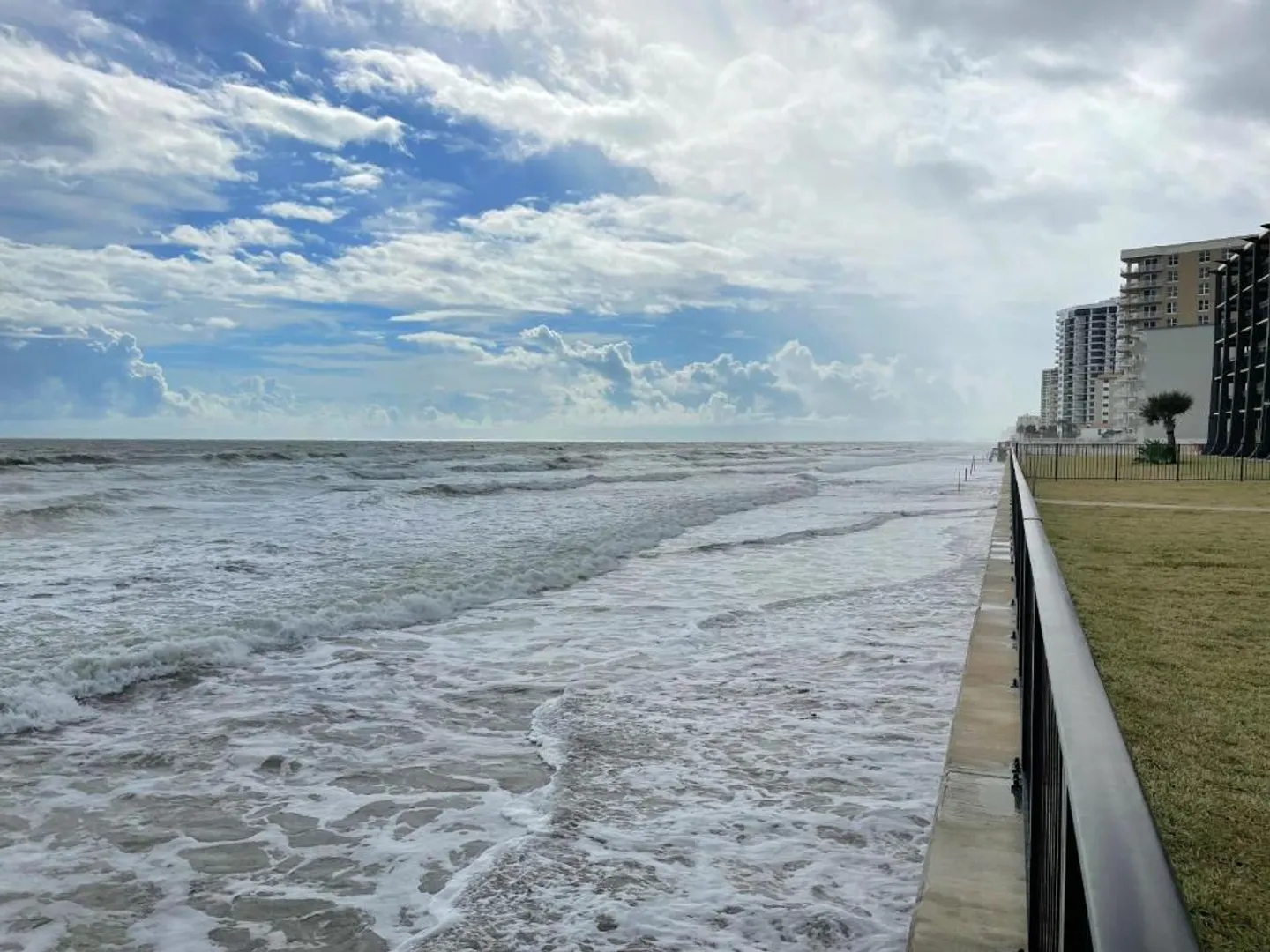 Ocean waves washing up against a seawall beside a grassy lawn and beachfront buildings under a cloudy sky.
