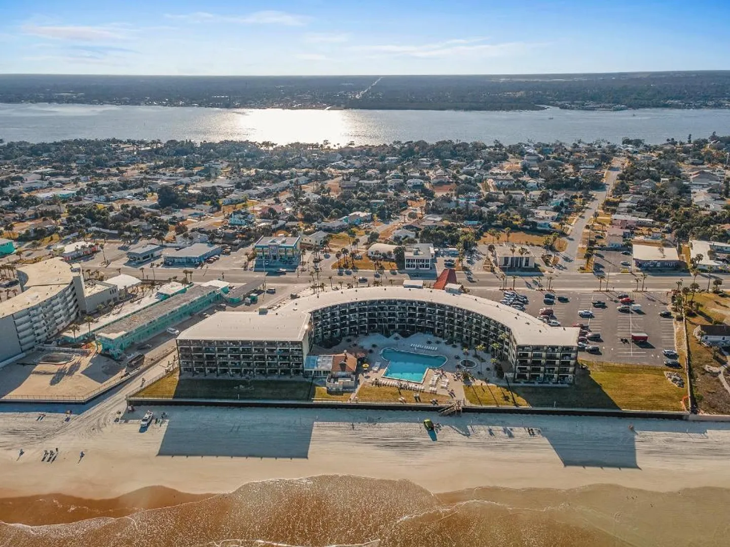 Aerial view of a curved beachfront condominium complex with an outdoor swimming pool, sandy beach, and surrounding coastal town.