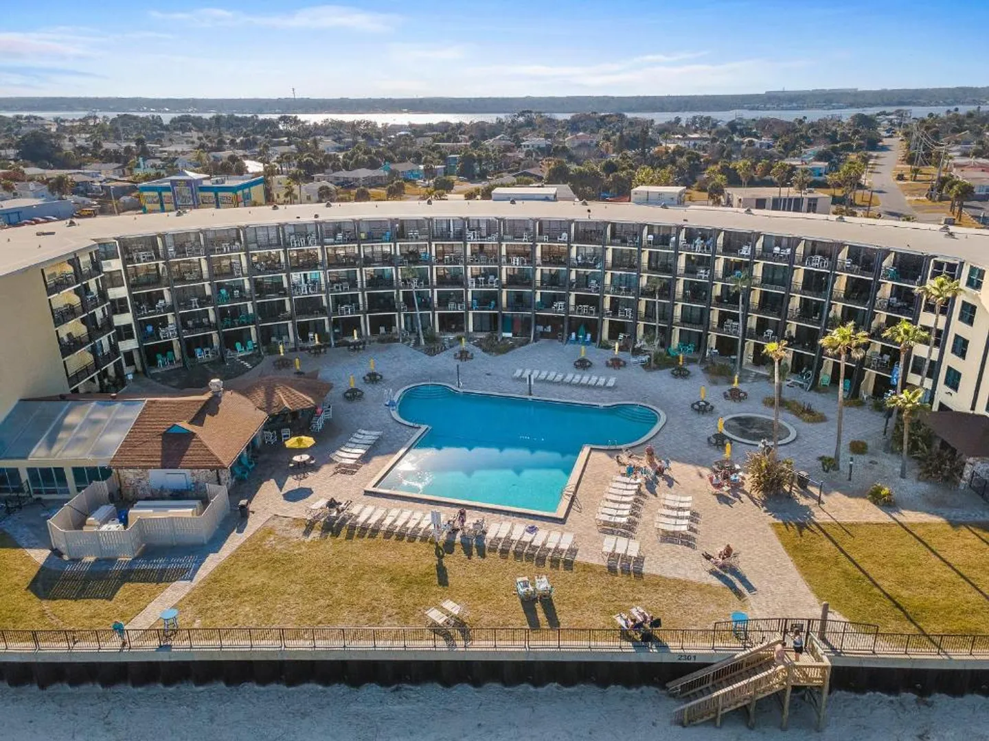 Aerial view of a curved multi-story condominium building featuring a swimming pool, sundeck with lounge chairs, and beach access.