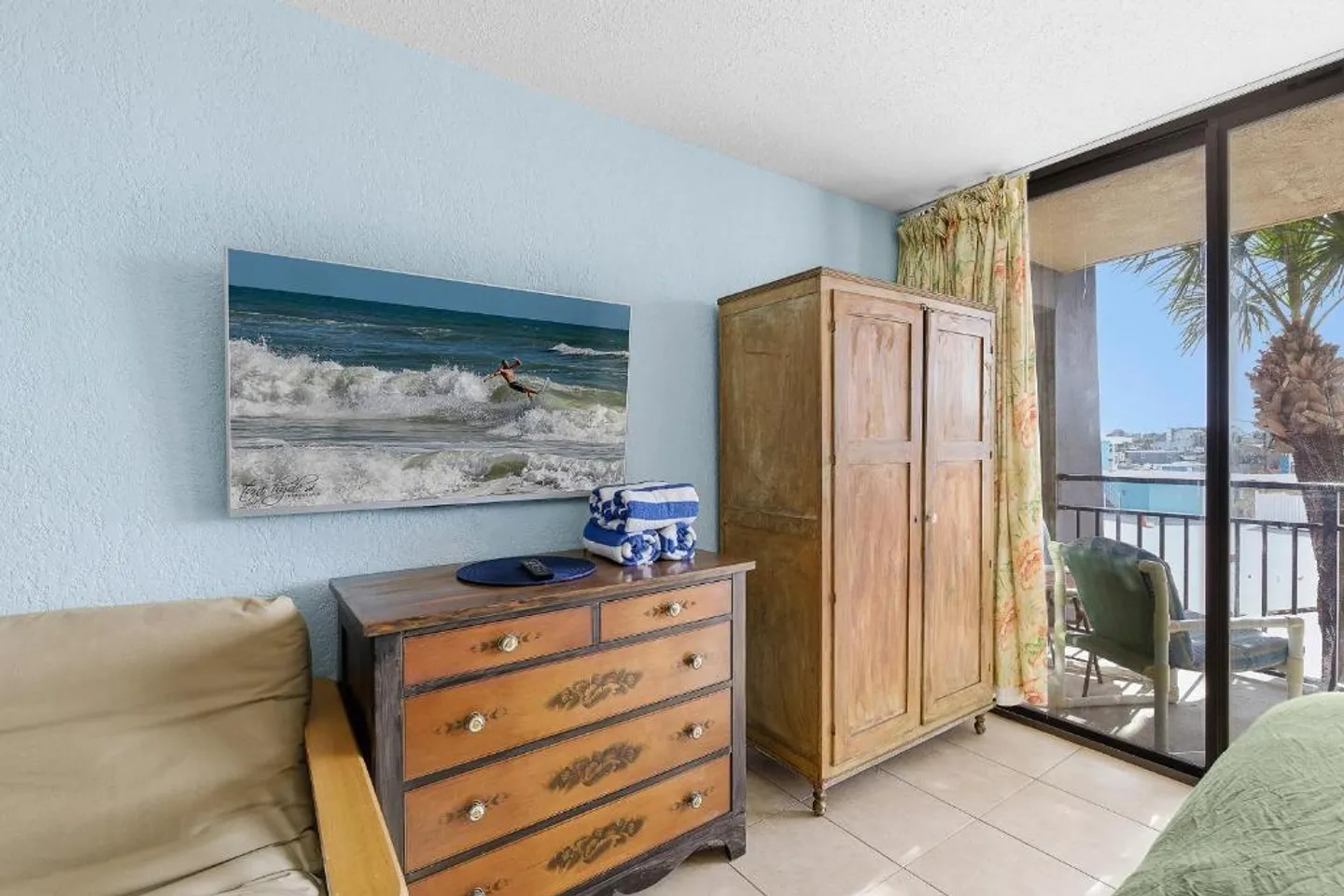Bedroom corner with light blue walls, a wooden dresser, a wardrobe armoire, and sliding glass doors leading to a balcony.