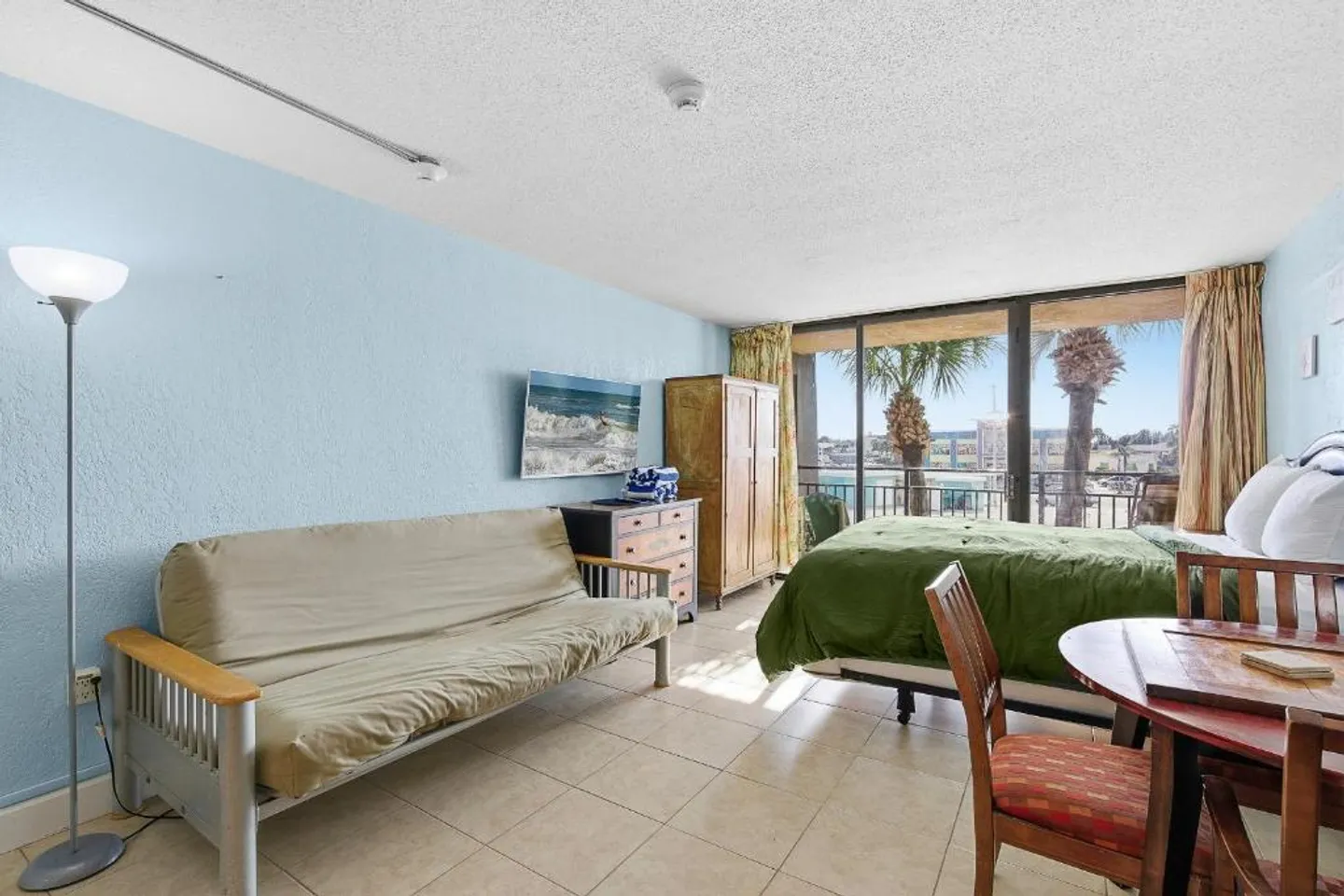 Studio bedroom featuring a bed, futon sofa, tiled floors, and sliding glass doors opening to a balcony with waterfront views.