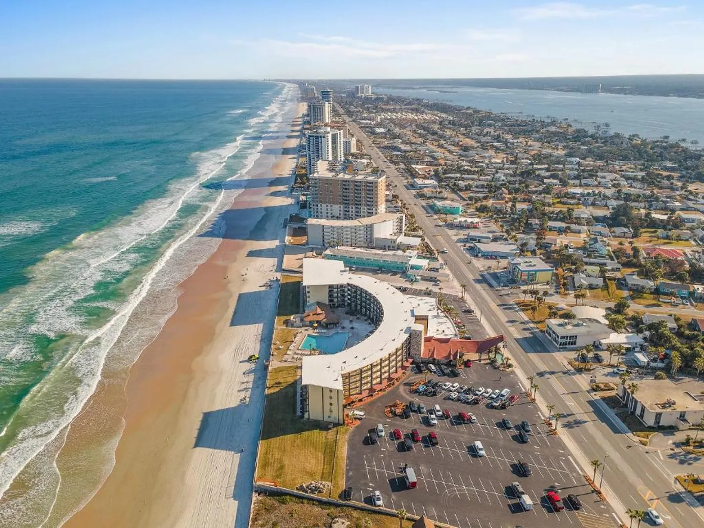Aerial view of a beachfront curved hotel building with a swimming pool, parking lot, sandy shoreline, and coastal skyline under a blue sky.